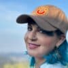 Capacete com sorriso amarelo bordado, mulher com cabelo azul e brincos estilo borboleta ao ar livre, sorriso e expressões modernas.