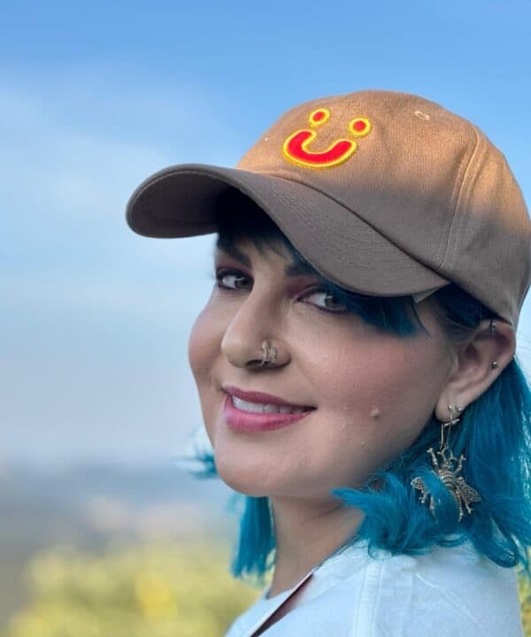 Capacete com sorriso amarelo bordado, mulher com cabelo azul e brincos estilo borboleta ao ar livre, sorriso e expressões modernas.