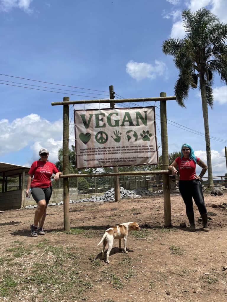 Alt: Pessoas com roupas veganas ao ar livre com cachorro, sinalização vegan na fazenda.